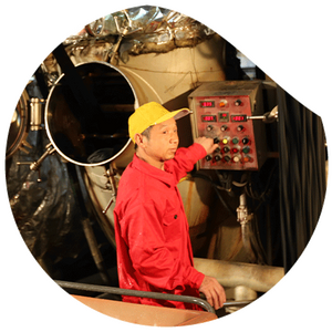 Ein Arbeiter in roter Arbeitskleidung und gelber Kappe bedient eine industrielle Steuerkonsole in einer Fabrikumgebung. Im Hintergrund ist ein großer metallischer Tank mit offener Luke zu sehen.